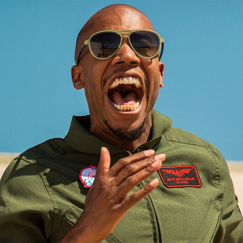 Un homme vêtu d’une combinaison verte de style militaire et de lunettes de soleil aviateur vertes cadet avec des verres dégradés verts crie joyeusement.