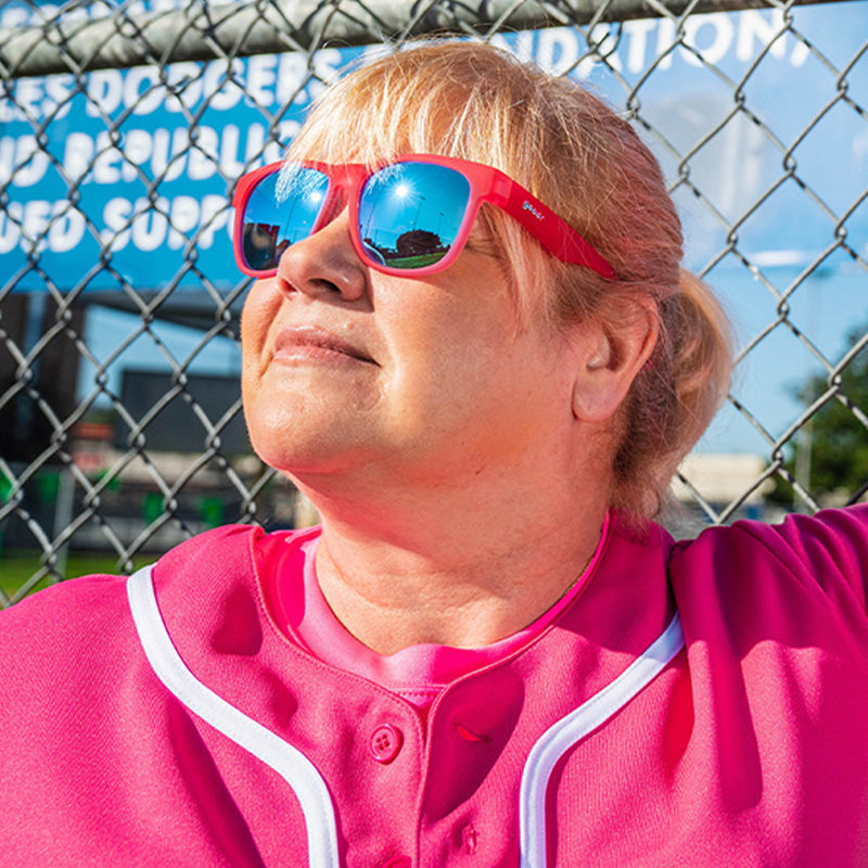 Une femme vêtue d'un uniforme de baseball rose vif et de lunettes de soleil rose vif à verres bleus s'appuie sur une clôture dans un rayon de soleil.