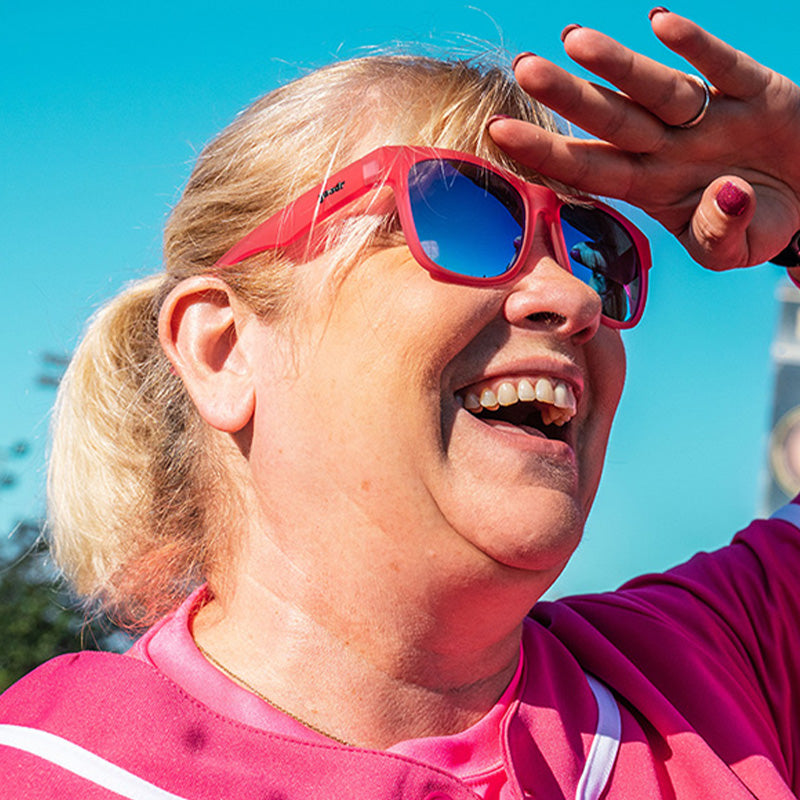 Vue de trois quarts d'angle d'une dame en uniforme de baseball rose et lunettes de soleil roses avec verres bleus, souriant sur un terrain de baseball.
