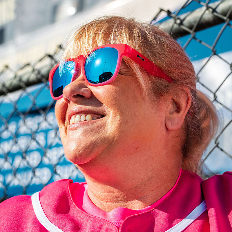 Une femme portant un uniforme de baseball rose et des lunettes de soleil roses avec des verres réfléchissants bleus sourit, adossée à une clôture.