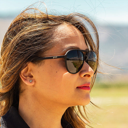 Une femme vêtue d'une combinaison noire et portant des lunettes de soleil aviateur noires avec des verres noirs non réfléchissants regarde férocement devant elle.