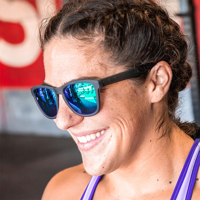 Une femme portant des lunettes de soleil grises avec des verres réfléchissants verts polarisés, souriant en regardant sur le côté.