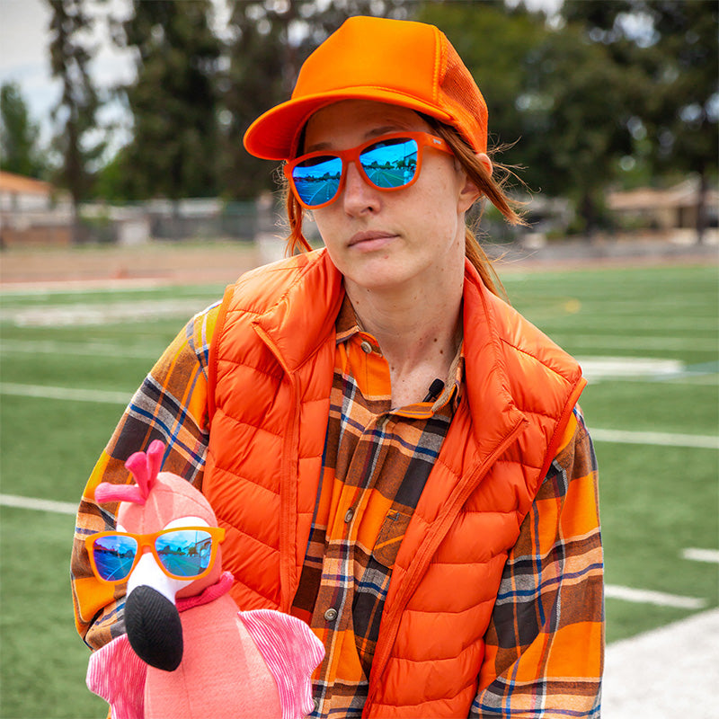 Une femme vêtue d'une tenue orange vif tient un flamant rose en peluche, toutes deux portant des lunettes de soleil orange à verres bleus.