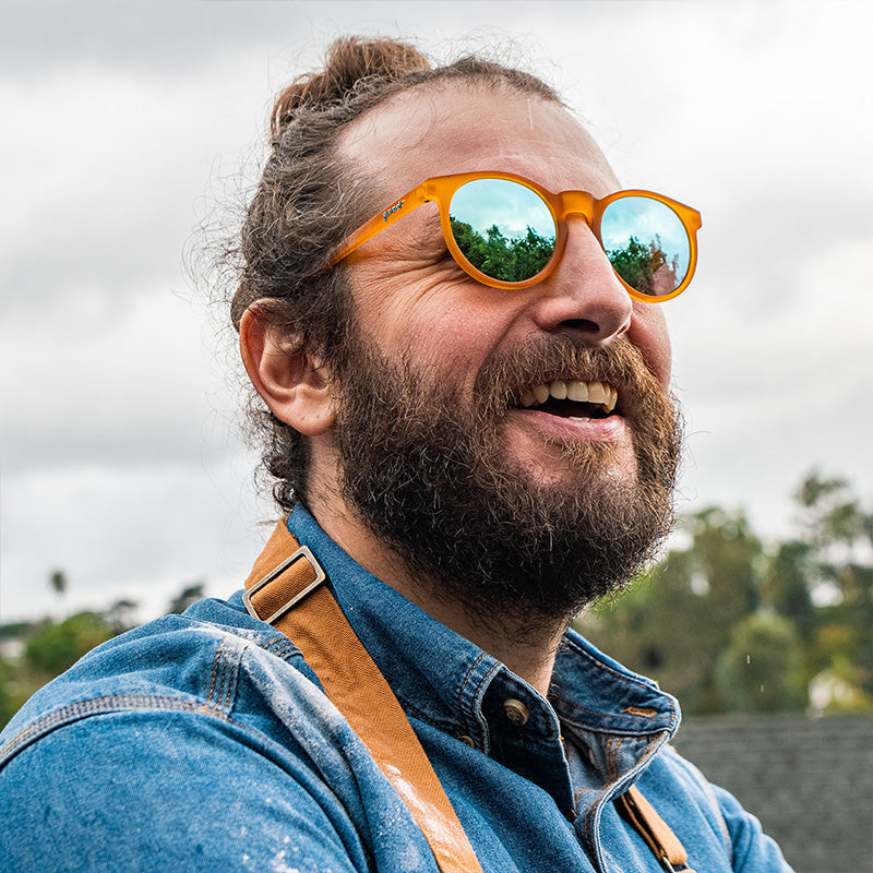 Un homme barbu avec un chignon d’homme sourit portant des lunettes de soleil rondes orange avec des verres bleu clair et un tablier recouvert de farine.