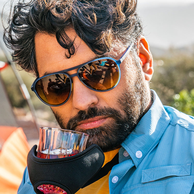 Un homme barbu portant des lunettes de soleil bleu aviateur renifle du whisky à l'aide d'un appareil qui tient le verre et que l'on pourrait voir dans un catalogue Skymall.