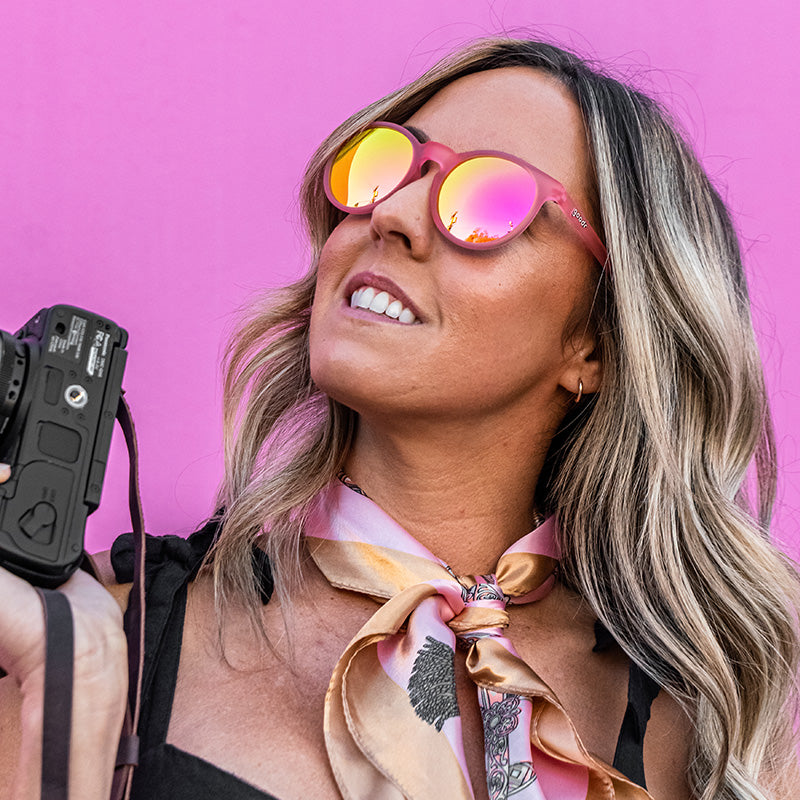 Une femme élégante portant des lunettes de soleil roses rondes et tenant un appareil photo regarde sur le côté, un mur rose vif derrière elle.