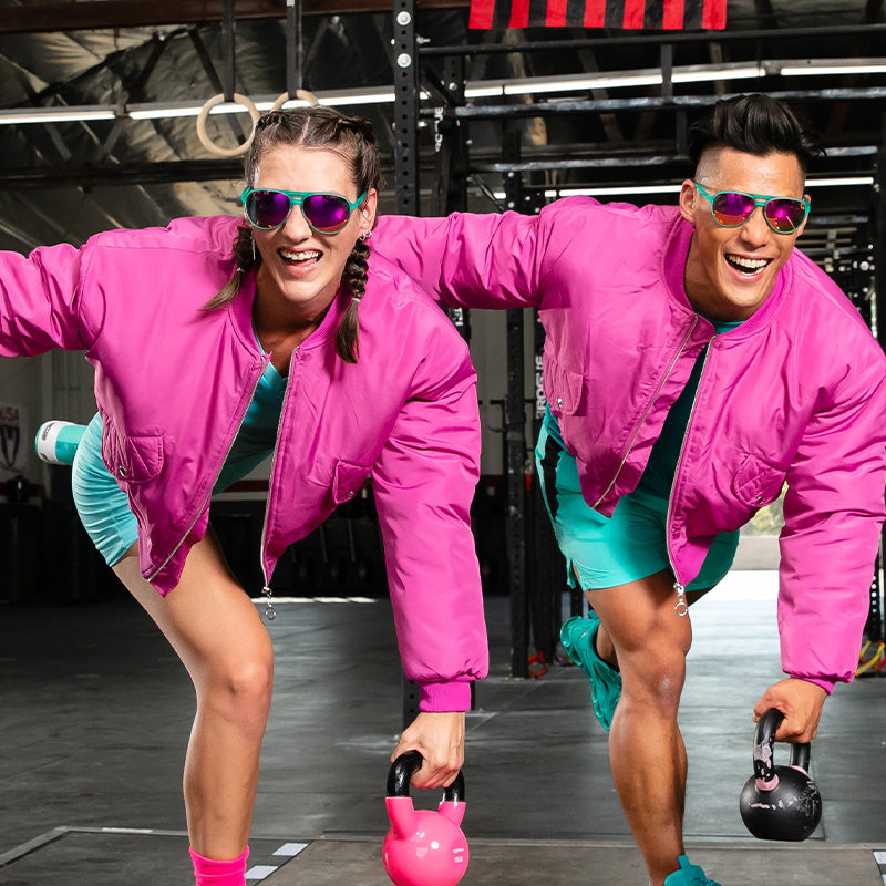 Un homme et une femme souriants vêtus de blousons d’aviateur roses et d’aviateurs turquoise avec des verres réfléchissants roses font une séance d’entraînement de kettlebell.