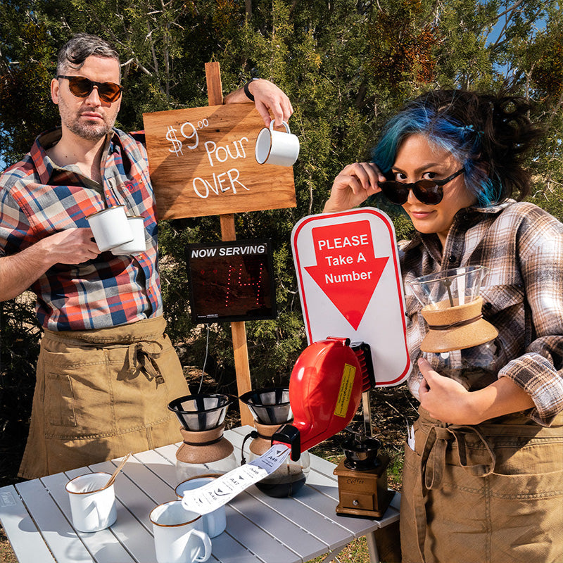 Deux baristas branchés portant des lunettes de soleil rondes aux verres bruns posent à un stand de café artisanal.
