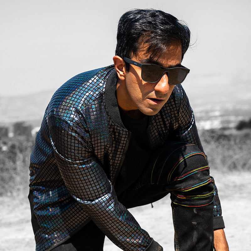 Un homme portant des lunettes de soleil noires à verre unique et une veste réfléchissante sombre et futuriste s'avance en regardant devant lui.
