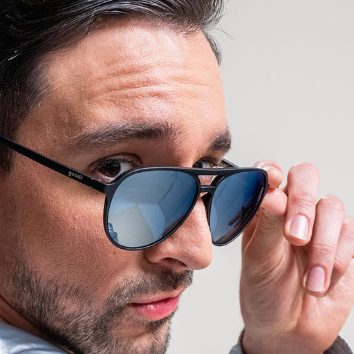 Un homme regarde par-dessus son épaule, des lunettes de soleil d'aviateur noires et chromées glissant sur son nez.