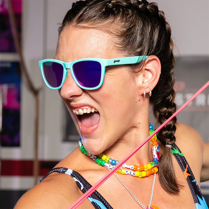 Une femme portant des colliers en plastique arc-en-ciel et des lunettes de soleil bleues avec des verres réfléchissants violets grogne férocement dans une salle de sport.