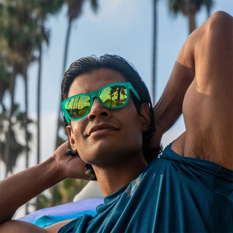 Un homme musclé et souriant porte des lunettes de soleil sarcelles avec des verres réfléchissants sarcelles et fléchit ses biceps, des palmiers au loin.