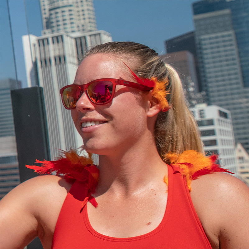 Une femme portant des lunettes de soleil rouges avec des verres rouges rehaussés de plumes rouges et orange, avec un horizon urbain derrière elle.