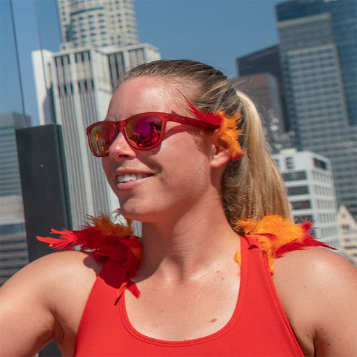 Une femme portant des lunettes de soleil rouges avec des verres rouges rehaussés de plumes rouges et orange, avec un horizon urbain derrière elle.