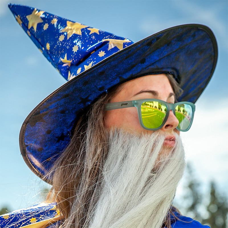Une femme portant un chapeau de sorcier et une grosse fausse barbe regarde dehors, portant des lunettes de soleil translucides bleu clair avec des verres réfléchissants dorés.