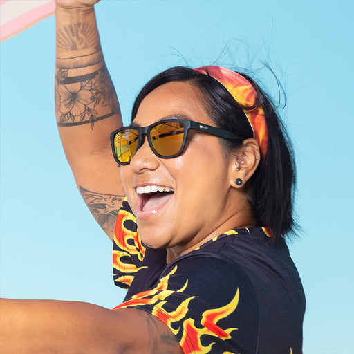 Une femme portant des lunettes de soleil noires avec des verres réfléchissants de couleur ambre, une chemise à motif de flammes et un bandeau rit.
